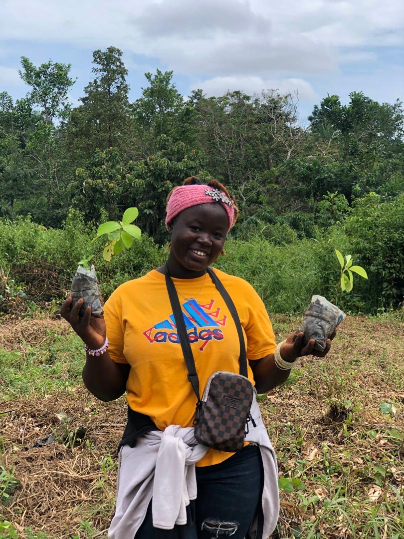 Smiling woman wearing a pink headband standing among young tree seedlings and forest, Sierra Leone