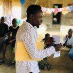 A man in a white patterned shirt stands and speaks while holding papers and a yellow folder, addressing a seated audience during a community meeting in a Sierra Leonean village. Attendees sit on a low ledge behind him, with colourful laundry hanging on a line in the background.