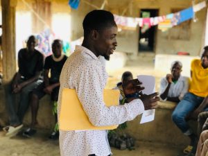 A man in a white patterned shirt stands and speaks while holding papers and a yellow folder, addressing a seated audience during a community meeting in a Sierra Leonean village. Attendees sit on a low ledge behind him, with colourful laundry hanging on a line in the background.