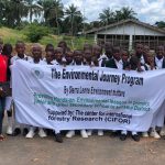 A group of students in school uniforms stand outdoors holding a large banner that reads "The Environmental Journey Program by Sierra Leone Environment Matters," describing hands-on environmental lessons in Kenema District schools supported by the Center for International Forestry Research (CIFOR), with palm trees and a building in the background.