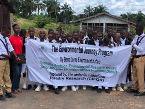 A group of students in school uniforms stand outdoors holding a large banner that reads "The Environmental Journey Program by Sierra Leone Environment Matters," describing hands-on environmental lessons in Kenema District schools supported by the Center for International Forestry Research (CIFOR), with palm trees and a building in the background.