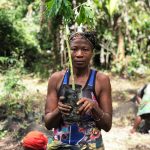 A woman wearing a patterned headwrap and blue tank top holds a tall young tree sapling growing in a black planting bag, standing in front of dense green forest and banana plants.