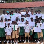 A large group of students in school uniforms pose outdoors in front of a building, proudly holding up their completed drawings and mind-map worksheets from an environmental education activity.