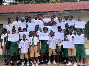 A large group of students in school uniforms pose outdoors in front of a building, proudly holding up their completed drawings and mind-map worksheets from an environmental education activity.
